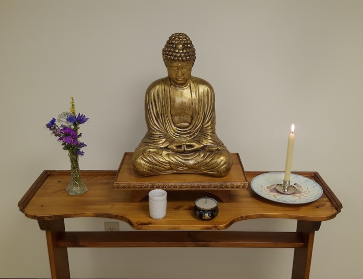 A photo of a Buddhist altar, consisting of a wooden platform with a gold-coloured Buddha statue, a lit candle, a small incense holder, a white teacup, and an arrangement of purple flowers in a narrow glass vase.