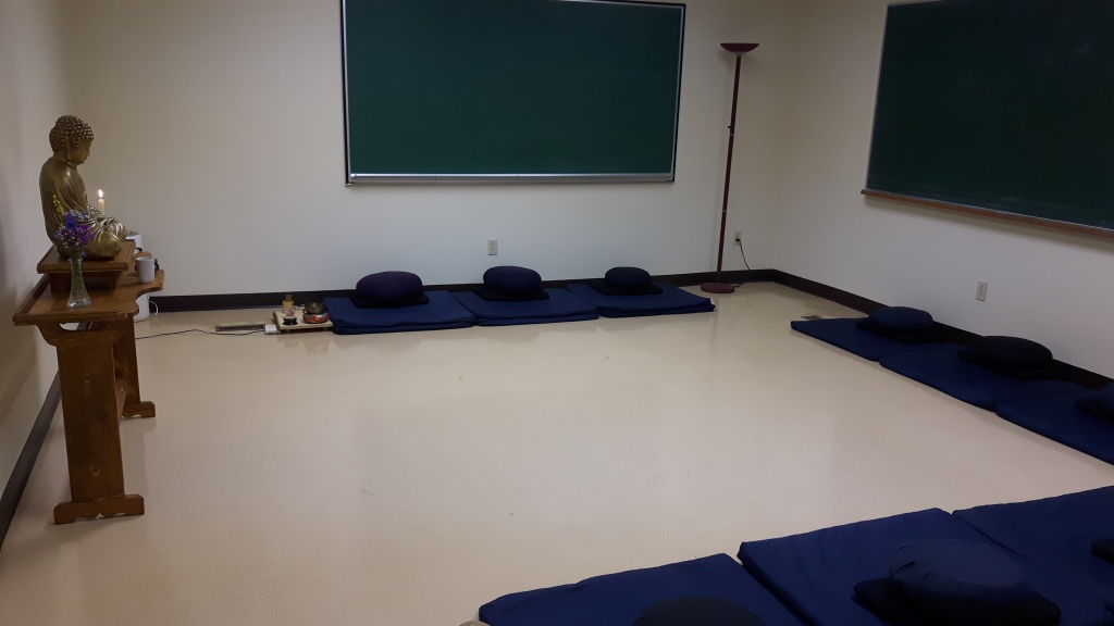 Photo of our meditation room, which shows a Buddhist altar against one wall, and nine dark blue sets of meditation cushions arranged against the three other walls.